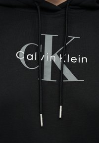 Felpa nera con cappuccio, logo "cK" grigio e scritta bianca "Calvin Klein" sul petto, con coulisse nere con punte argento.