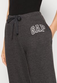 Person, der dunkelgraue Jogginghosen mit schwarzem Kordelzug und weißem "GAP"-Logo auf dem Oberschenkel trägt, mit einer Hand teilweise in der Tasche.