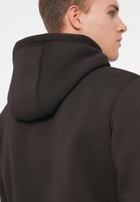 Felpa cappuccio marrone scuro con una texture morbida, caratterizzata da un cappuccio classico e dettagli di cucitura a vista, progettata per una vestibilità comoda.