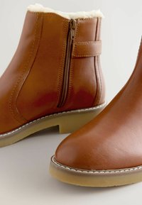 Bruine leren enkellaarzen met zijrits en witte stiksels, voorzien van een lichtbeige zool en een zachte crèmekleurige binnenvoering.