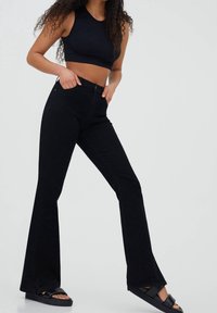 Jeans de campana negros con una textura suave, cintura alta y bolsillos frontales, combinados con una camiseta corta negra y sandalias planas negras.