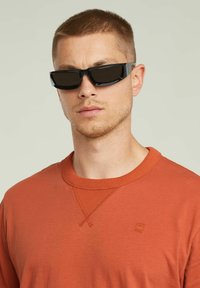 Occhiali da sole neri a forma rettangolare con lenti oscurate, indossati da una persona in una maglietta a maniche lunghe arancione con colletto texturizzato e cuciture.