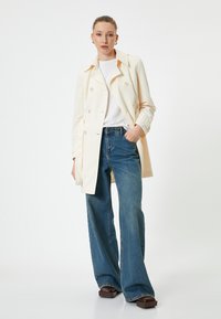 Cappotto trench doppio petto color crema con un grande colletto, abbinato a jeans blu a vita alta e gamba larga e scarpe con platform color marrone scuro.