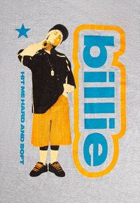 Grå t-skjorte med et grafisk trykk av en person i sort skjorte og oransje shorts, med blå og oransje tekst som lyder "billie" og "HIT ME HARD AND SOFT."