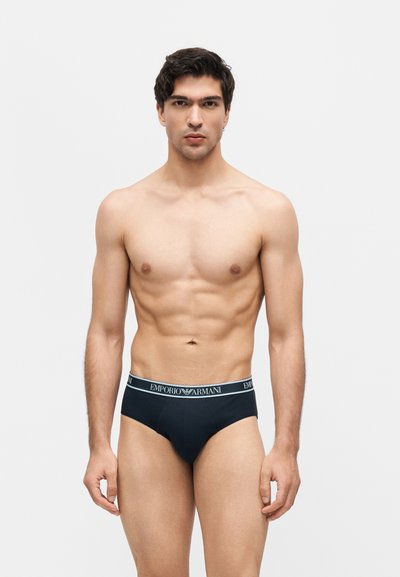 Άνδρας μοντέλο με σκούρα μαλλιά που φοράει μαύρα εσώρουχα Emporio Armani, στέκεται μπροστά σε ένα απλό ανοιχτό γκρι φόντο, κοιτάζοντας μπροστά με ουδέτερη έκφραση.
