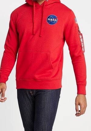 Czerwona bluza z kapturem z logo NASA na piersi, kieszenią na zamek i naszywką amerykańskiej flagi na lewym rękawie, noszona z ciemnoniebieskimi dżinsami.