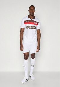 Biely futbalový dres s červeným a hnedým pruhovaným vzorom, krátkymi rukávmi, doplnený o biele šortky a ponožky. Obsahuje logo Puma a značky sponzora.