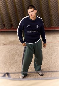 Ένας νέος άνδρας με κοντά μαλλιά στέκεται έξω φορώντας έναν ναυτικό πουλόβερ με λευκές ρίγες, πράσινα joggers και παπούτσια με σχέδια.