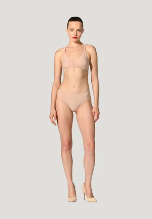 Frau trägt nudefarbenen BH und Slip mit beigen High Heels, steht vor schlichtem hellgrauem Hintergrund.