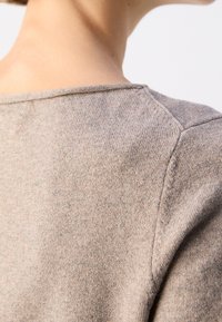 Gros plan sur le haut du dos et l'épaule d'une personne portant un pull en maille marron clair avec des détails de couture visibles.