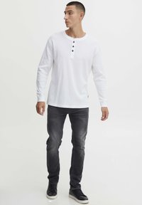 Witte lange mouwen henley-shirt met drie zwarte knopen, gecombineerd met donkergrijze slim-fit jeans en zwarte sneakers. Glad stoftexture.