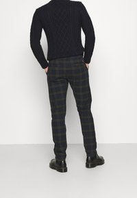 Maglione navy a maglia grossa abbinato a pantaloni a quadri in verde scuro e blu. I pantaloni hanno una vestibilità attillata e due tasche posteriori. Stivali neri.