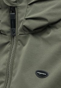 Close-up van olijfgroen jasstof met een zigzagpatroon, voorrits en zwarte ovale Raquwear-logo patch.