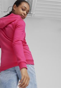 Svijetloružičasta dukserica s kapuljačom i vezicom, u kombinaciji s svijetloplavim trapericama. Jednostavan, ležeran dizajn s glatkom teksturom.