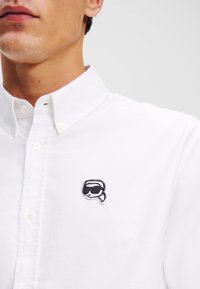 Weißes Hemd mit Kragen und Knopfleiste, mit einem schwarzen Stickdesign auf der linken Brust. Der Stoff wirkt glatt und leicht.