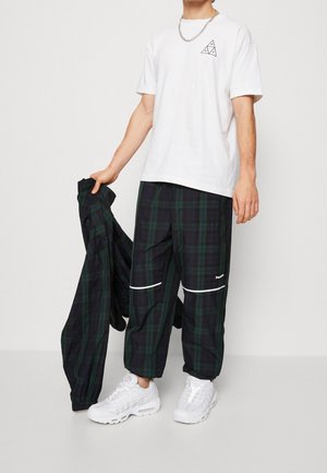 Mies, joka pukeutunut valkoiseen T-paitaan, vihreisiin ja tummansinisiin ruudullisiin housuihin, pitää kädessään samansävyistä ruudullista takkia ja valkoisia tennareita, yksiväristä taustaa vasten.