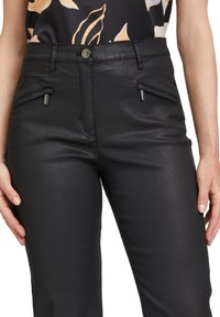 Schwarze Kunstlederhose mit glatter Textur, ausgestattet mit zwei Frontreißverschlusstaschen und einer figurnahen Passform. Metallische Akzente an den Taschen.
