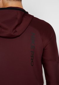 Bordeaux Nike Pro hættetrøje med en tekstureret stof og ribbede detaljer, prydet med et sort logo på siden og en tætsiddende hætte.
