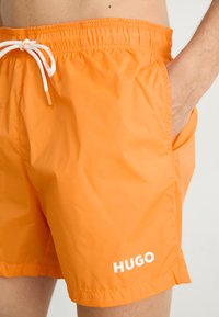 Oranžové plavky s elastickým pásom, šnúrkou na sťahovanie, bočnými vreckami a bielym logom "HUGO" na dolnej ľavej nohe. Hladká textúra.