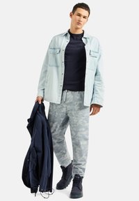 Camicia in denim azzurro chiaro sopra un maglione blu navy, pantaloni jogger grigi con motivo camouflage e scarpe alte nere, mentre tiene un giubbotto blu navy. Outfit fresco e informale.