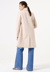 Abrigo de mezcla de lana beige con un acabado texturizado, que cuenta con un cuello con muesca, un corte ajustado y un dobladillo trasero partido, combinado con jeans azules acampanados.