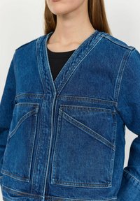 Jeans jakna v temno modri barvi z V-izrezom, dvema velikima sprednjima žepoma, kontrastnim šivom in teksturiranim zaključkom.