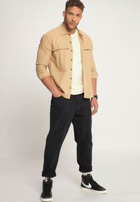 Camisa beige de botones con dos bolsillos en el pecho, usada sobre una camiseta blanca. Pantalones negros arremangados y zapatillas altas negras.