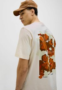 Cream-colored t-shirt featuring a floral graphic print in orange, brown, and green on the back, with "Creative Workshop" text at the bottom. Brown cap.