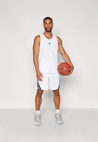 Valkoinen urheilullinen hihaton paita mustalla logolla, valkoiset koripalloshortsit mustilla sivuraidoilla, vaaleat lenkkarit, pitelee koripalloa.