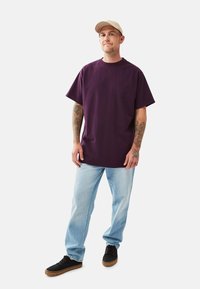 Mann mit beiger Mütze, lila oversized T-Shirt, hellblauen Jeans, schwarzen Sneakers und sichtbaren Armtattoos, der vor weißem Hintergrund steht.