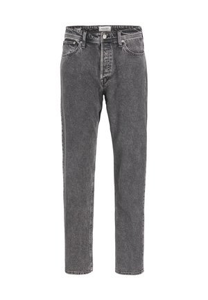 Person iført en enkel hvid T-shirt med korte ærmer, løse grå syrevaskede jeans og hvide sneakers med sorte detaljer, stående mod en ensfarvet baggrund.