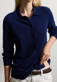 Cardigan en laine bleu marine avec un devant à boutons, un col pointu, des poignets côtelés et une texture douce, porté sur un pantalon blanc et une ceinture marron.