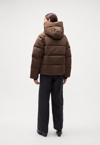 Brun pufferjakke med hætte, der har et quiltet design og en boxy form. Steret med mørke vidde bukser og hælede støvler.
