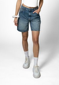Denimshorts i mellomblått med løs passform, oppbrettede falmer og frontlommer. Brukt med hvite høye sneakers og lyse sokker.