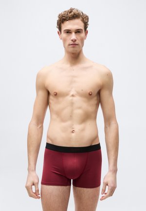 Νέος άντρας με σγουρά μαλλιά που στέκεται, φοράει κόκκινα μποξεράκια με μαύρη μέση, ουδέτερη έκφραση προσώπου, ανοιχτό φόντο.