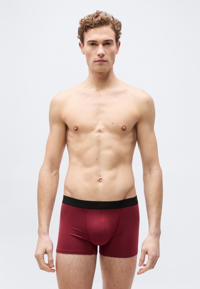 Νέος άντρας με σγουρά μαλλιά που στέκεται, φοράει κόκκινα μποξεράκια με μαύρη μέση, ουδέτερη έκφραση προσώπου, ανοιχτό φόντο.