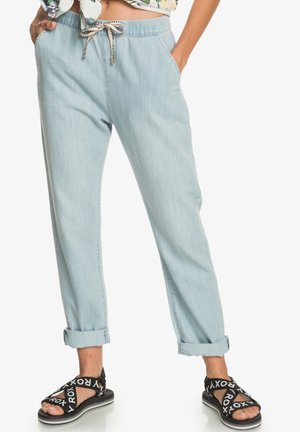 Personne portant un pantalon bleu clair retroussé avec une taille à cordon et des sandales noires avec des brides blanches "ROXY", debout sur fond blanc.