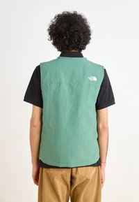 Green vest with a smooth texture, round neckline, and a small white logo on the upper back, worn over a black short-sleeve shirt.