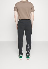 Una maglietta marrone a maniche corte abbinata a pantaloni sportivi neri con il logo Nike bianco; include sneakers verdi e bianche con un design testurizzato.