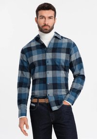 Camisa de franela a cuadros en tonos de azul y gris, con mangas largas, botones en la parte delantera y un bolsillo en el pecho. Llevada sobre un suéter de cuello alto blanco.