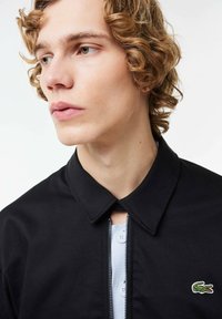 Melns rāvējslēdzēja mētelis ar izteiktu apkakli, kuram kreisajā pusē ir neliels zaļš krokodila simbols un gaiši zils iekšējais slānis ar pogām.