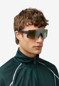 Akiniai nuo saulės su kampuotais, stambiais rėmeliais; juodas su taškučiais rėmas; žalių atspalvių lęšiai; papildyti aukštu apykakle žalia striuke.