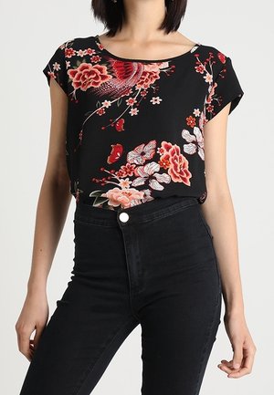 Kobieta w czarnej bluzce z krótkim rękawem w kwiatowy wzór, włożonej do czarnych, wysokiej talii dżinsów, pozująca z rękami swobodnie opuszczonymi wzdłuż ciała.