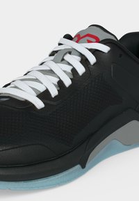 Μαύρο αθλητικό παπούτσι με λευκά κορδόνια, γκρι γλώσσα με κόκκινο λογότυπο, επάνω μέρος από δίχτυ και γαλάζια και γκρι σόλα ορατή σε λευκό φόντο.