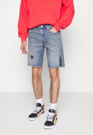 Lysblå denimshorts med frynset kant, udstyret med en lap og slidte detaljer. Båret sammen med en rød hættetrøje og sorte sneakers.
