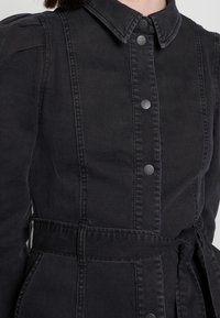 Robe chemise en denim noir avec un col, fermeture à boutons sur le devant, ceinture à la taille et détails de coutures subtiles sur les côtés.
