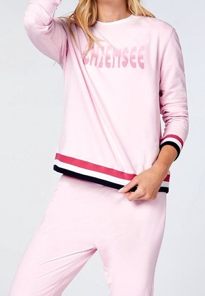 Kvinde iført en lyserød sweatshirt med teksten "CHIEMSEE", med røde, hvide og sorte stribede manchetter og kant, kombineret med matchende bukser.