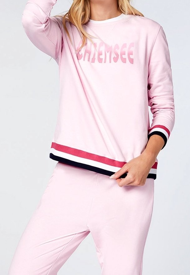 Femme portant un sweat-shirt rose clair avec le texte "CHIEMSEE", avec des poignets et un bas rayés rouge, blanc et noir, assorti à un pantalon.