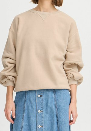 Person iført beige oversized sweatshirt med opsamlede ærmer og højtaljet blå denim nederdel med knapper foran mod hvid baggrund.