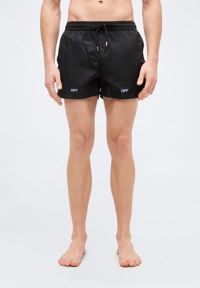 Homme portant un short de bain noir avec le texte « OFF » sur chaque jambe, debout pieds nus devant un fond clair uni.